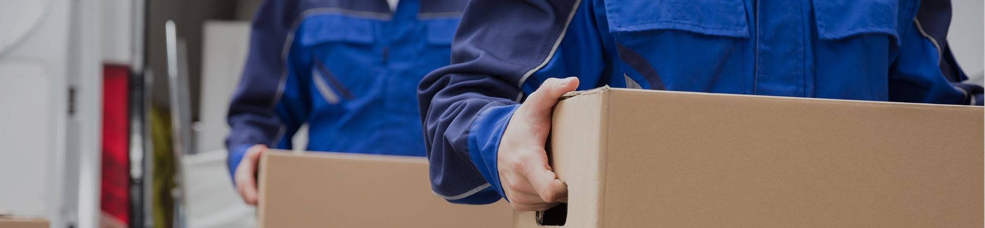 Zwei Personen in blauer Arbeitskleidung tragen Kartons, nur Oberkörper und Hände sichtbar.